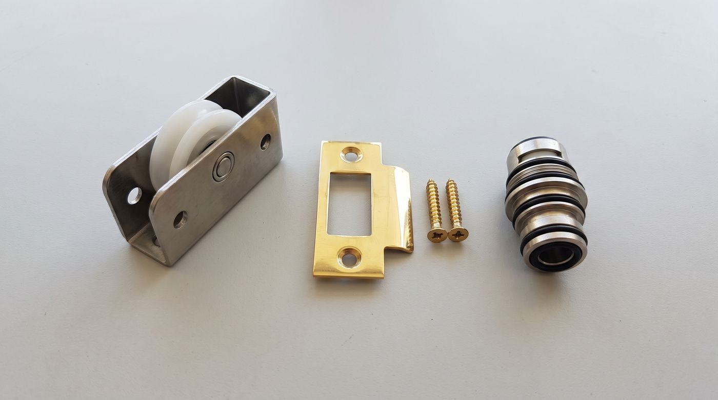 A new sliding door roller in its steel housing, a brass strike plate with two screws and a faucet cartridge with black o-rings, laid out on a plain gray surface